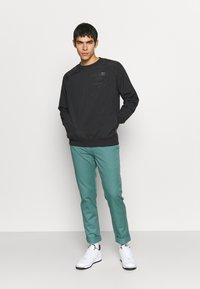 Svart hettegenser med teksturdetaljer og logo foran, sammen med teal slim-fit bukser og hvite joggesko. Plassert mot en ensfarget bakgrunn.