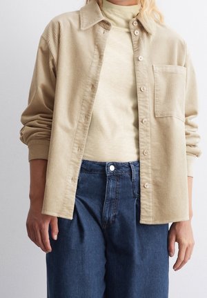 Beżowa koszula sztruksowa na guziki, noszona rozpięta na kremowym golfie, zestawiona z wysokim stanem niebieskich dżinsów.