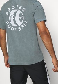 Camiseta de algodón gris con un gran gráfico blanco que dice “PROPER FOOTBALL” y un diseño circular de balón de fútbol en la parte trasera. Mangas cortas, ajuste casual.