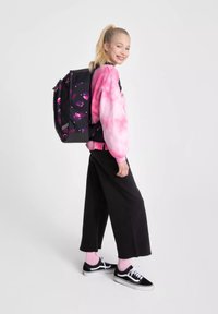 Jeune personne avec des cheveux blonds en queue de cheval portant un sweat-shirt rose tie-dye, un pantalon large noir et des baskets noires avec un sac à dos fleuri.