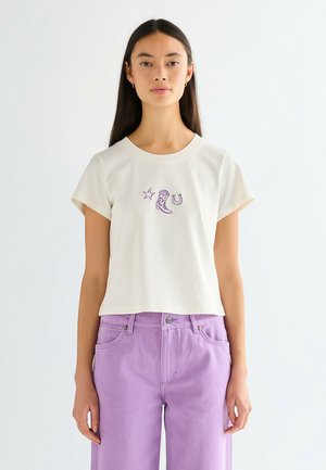 Młoda kobieta o długich ciemnych włosach, ubrana w białą koszulkę z fioletowymi wzorami kowbojskich butów, gwiazdy i podkowy, do tego fioletowe dżinsy.