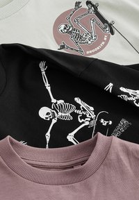 Drei T-Shirts: eines grau mit einem grafischen Motiv eines kreisförmigen Skeletts auf Skateboard, eines schwarz mit einem Skelettdesign, eines pink mit einem texturierten Kragen.