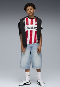 Pojke står klädd i en röd och vit randig PSV Eindhoven fotbollströja, ljus denimshorts, vita Puma-strumpor och sneakers, svart sweatshirt över axlarna.