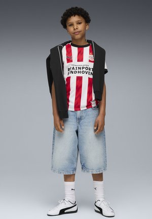 Ragazzo in piedi che indossa una maglia da calcio a strisce rosse e bianche del PSV Eindhoven, pantaloni corti in denim chiaro, calze bianche Puma e scarpe da ginnastica, felpa nera sulle spalle.