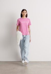 Rozā kokvilnas T-krekls ar "Calvin Klein Jeans" logotipu, apvienots ar gaiši zili vaļīgiem džinsiem un baltām sporta kedām, turot baltu džemperi.