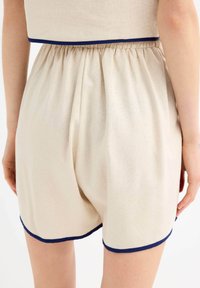 Pantalones cortos beige con una tela texturizada, cinturilla elástica y ribete azul marino en el dobladillo. Pliegues visibles en la cinturilla.