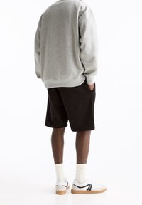 Persona che indossa un felpa grigia, pantaloni neri, calze bianche e sneakers bianche con suola in gomma, in piedi con le mani in tasca girata di spalle.