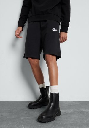 Osoba ubrana w czarne spodenki Nike, czarne masywne buty, białe skarpetki oraz czarną bluzkę z długim rękawem, stojąca przy białej ścianie.