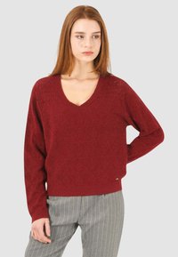 Maglione borgogna a maglia con scollo a V, motivo geometrico testurizzato, maniche lunghe e un piccolo marchio sull'orlo. Accoppiato con pantaloni a righe.