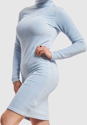 Frau trägt ein hellblaues, geripptes Strickkleid mit Rollkragen und langen Ärmeln, posiert mit einer Hand in der Hüfte vor einfarbigem Hintergrund.