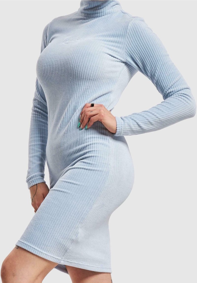 Femme portant une robe en maille côtelée bleu clair à col roulé avec des manches longues, posant avec une main sur la hanche devant un fond uni.