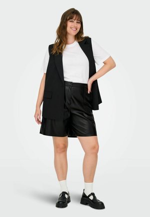 Blazer senza maniche nero sopra una maglietta bianca, abbinato a pantaloni corti in finta pelle nera e scarpe nere chunky, con calzini bianchi.