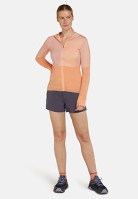Chaqueta ligera con cremallera en tonos naranjas, con capucha, mangas largas y agujeros para los pulgares; combinada con pantalones cortos oscuros y zapatillas deportivas negras.
