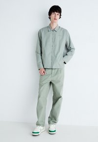 Jachetă verde deschis cu fermoar și guler, purtată cu pantaloni asortați și adidași albi cu accente verzi. Design minimalist.