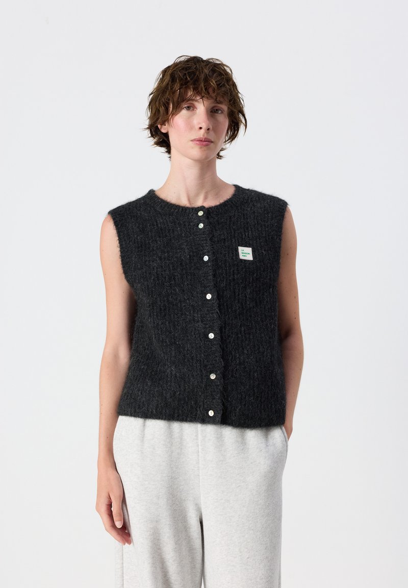 Persoon draagt een mouwloos zwart pluizig vest met knopen en een klein borstlabel en lichtgrijze broek tegen een effen achtergrond.