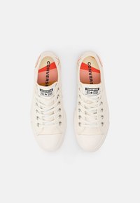 Beiga canvas sneakers med vita snören, rund tås och metallhål. Har en röd och orange accent på insulan och logotypen.
