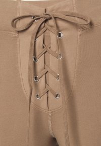 Hnědé, ribbované kalhoty s předním šněrováním a kovovými očky. Měkká tkanina s přiléhavým střihem a viditelným šitím.