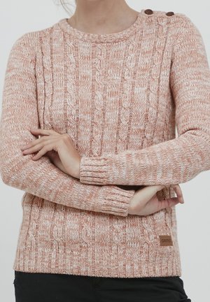 Femme portant un pull en maille torsadée rose avec des détails à boutons sur l'épaule, les bras croisés sur le torse, debout devant un fond uni.