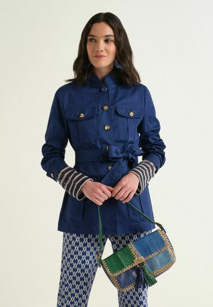 Frau trägt eine gebundene marineblaue Jacke über gestreiften Ärmeln, blau-weiß gemusterte Hose, hält eine grüne und blaue Patchwork-Handtasche.