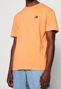 Krátký rukáv, oranžové bavlněné tričko s malou vícebarevnou nášivkou na hrudi. Volný střih se rovním lemem.