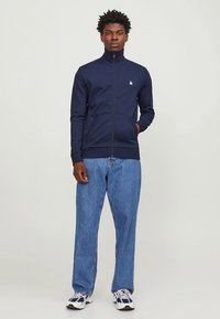Felpa con zip blu navy in tessuto morbido, con collo alto e due tasche frontali, abbinata a jeans a gamba dritta azzurri.