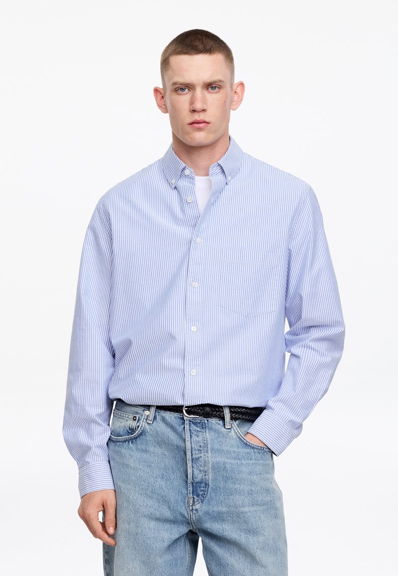 Jeune homme aux cheveux courts portant une chemise à boutons rayée bleu clair et blanc par-dessus un tee-shirt blanc et un jean bleu clair, debout avec une main dans la poche.