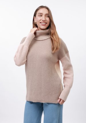 Pull en maille beige à col roulé avec une texture côtelée, épaules tombantes et manches longues. Présente un dégradé de couleur vers une tonalité rose plus claire.