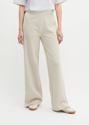 Άτομο με μπεζ παντελόνες φαρδιάς γραμμής, σε συνδυασμό με λευκά αθλητικά παπούτσια και άσπρη φαρδιά μπλούζα, στέκεται μπροστά σε απλό φωτεινό φόντο.