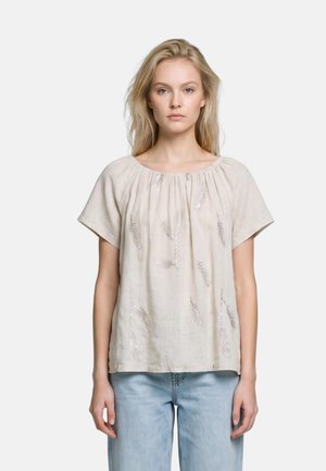 Giovane donna con lunghi capelli biondi che indossa una camicetta beige chiaro con motivi a foglie e jeans azzurri chiari, in piedi davanti a uno sfondo semplice.