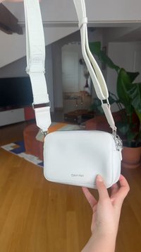 Λευκή τσάντα Calvin Klein crossbody με ρυθμιζόμενο λουράκι, κρατημένη από χέρι σε σαλόνι με φυτά και έπιπλα στο φόντο.