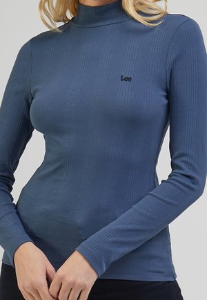 Femme portant un haut bleu côtelé ajusté à manches longues et col roulé avec un petit logo Lee noir sur la poitrine.