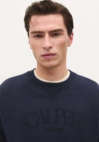 Marineblauwe sweatshirt met een ribgebreide halslijn en raglanmouwen. Voorzien van reliëftekst "SCALPERS THEATRE" op de borst.