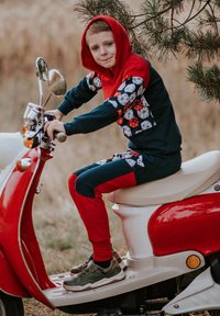 Czerwony i granatowy dres dziecięcy z wzorem w kości, noszony przez dziecko na czerwonej hulajnodze. Posiada kaptur i kontrastujące rękawy.