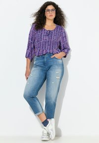 Violetti kuvioitu paita, jossa on 3/4-hihat, yhdistetty vaaleanpesuihin revittyihin farkkuihin ja hopeisiin lenkkareihin. Paidassa on käännetyt hihansuut ja sivutaskut.