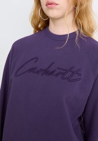 Purple crewneck sweatshirt features raised embroidery of "Carhartt" across the chest, made from a smooth cotton blend fabric.