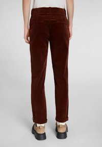 Henkilö, joka on pukeutunut tummanruskeisiin corduroy-housuihin, joissa on käänteet, vaaleisiin kenkiin mustilla pohjilla ja valkoisiin sukkiin, seisoo tasaisen taustan edessä.