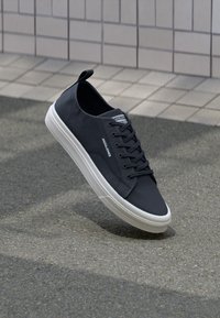 Schwarze Low-Top Sneaker mit weißer Sohle und Schnürsenkeln, markiert mit "Jack & Jones", schwebend über texturiertem Boden im Freien nahe einer gefliesten Wand.