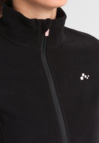 Close-up van een persoon die een zwarte fleecejack draagt met een hoge kraag en rits, met een klein wit logo op de borst.