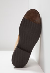 Suela de un zapato de ante beige con una parte inferior de goma negra texturizada y un detalle de cuero marrón cerca de la punta, en ángulo sobre una superficie blanca.