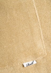 Beige Handtuch mit einer weichen, strukturierten Oberfläche und einer gestickten Kante. Ein kleines Etikett mit "Riell" ist in der unteren Ecke sichtbar.