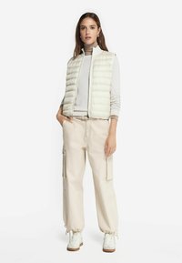 Licht gewatteerd, romig beige mouwloos vest over een lichtgrijze top, gecombineerd met losse, crèmekleurige cargobroeken en witte sneakers.