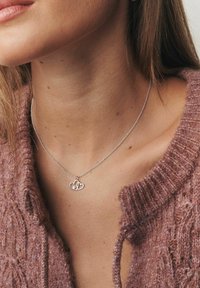 Collana d'argento con ciondolo a doppio cuore, caratterizzata da un piccolo diamante, indossata su un maglione lavorato a maglia color malva.