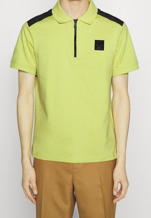 Mężczyzna ubrany w limonkowo-zieloną krótką koszulkę polo z czarnymi naszywkami na ramionach, czarnym zamkiem z przodu i brązowe spodnie, przedstawiony od szyi do bioder.