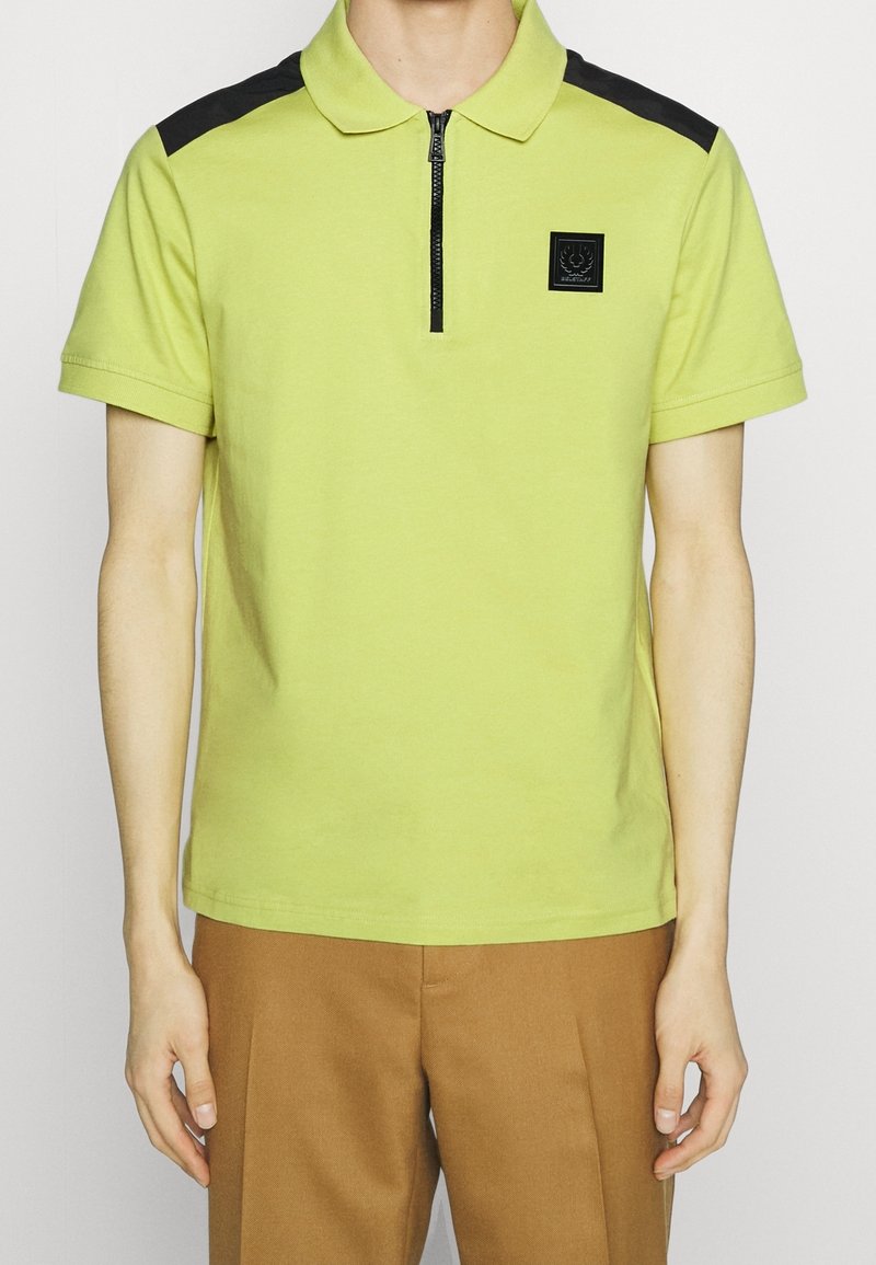Homme portant un polo manches courtes vert citron avec des empiècements noirs sur les épaules, une fermeture éclair noire à l'avant et un pantalon marron, visible du cou aux hanches.