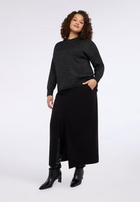 Maglione nero glitterato con vestibilità comoda, abbinato a una gonna lunga nera con spacco laterale. Il modello indossa stivaletti neri alla caviglia.