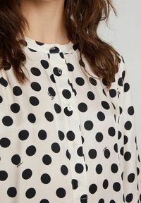 Blusa a pois bianchi con pois neri, dotata di colletto, chiusura con bottoni e un tessuto strutturato. Include piccole cuciture decorative.