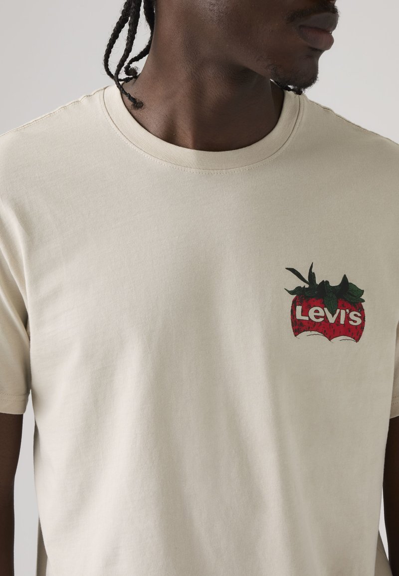 Hombre que lleva una camiseta beige de Levi's con un logotipo de fresa rojo y verde en el pecho izquierdo, mirando hacia la derecha con el cabello trenzado.