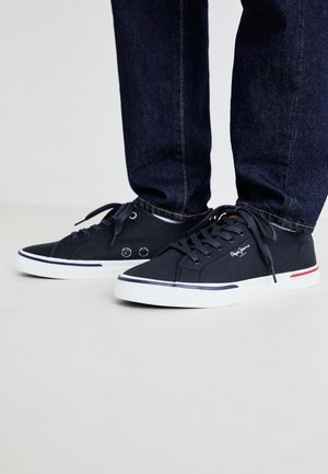 Mornarice platnene tenisice s bijelim potplatima, s okruglim vezicama i detaljem logotipa. Teksturirana tkanina s kontrastnim crvenim i plavim akcentima sa strane.