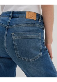 Blue denim jeans with a fitted design, featuring back pockets and a tan leather patch on the waistband displaying a logo.