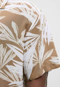 Chemise à manches courtes beige avec des motifs de feuilles blanches. Texture lisse, design avec col et coupe décontractée avec un détail de couture visible.